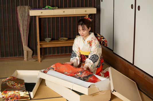 A Japanese woman in a floral kimono carefully unpacks a vibrant red kimono from a wooden box in a traditional tatami mat room.